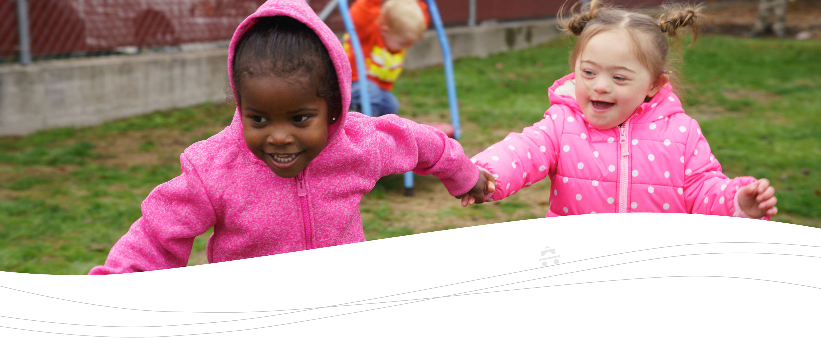 Two preschoolers hold hands while running outside.  Another preschooler is in the background jumping on a child size trampoline.